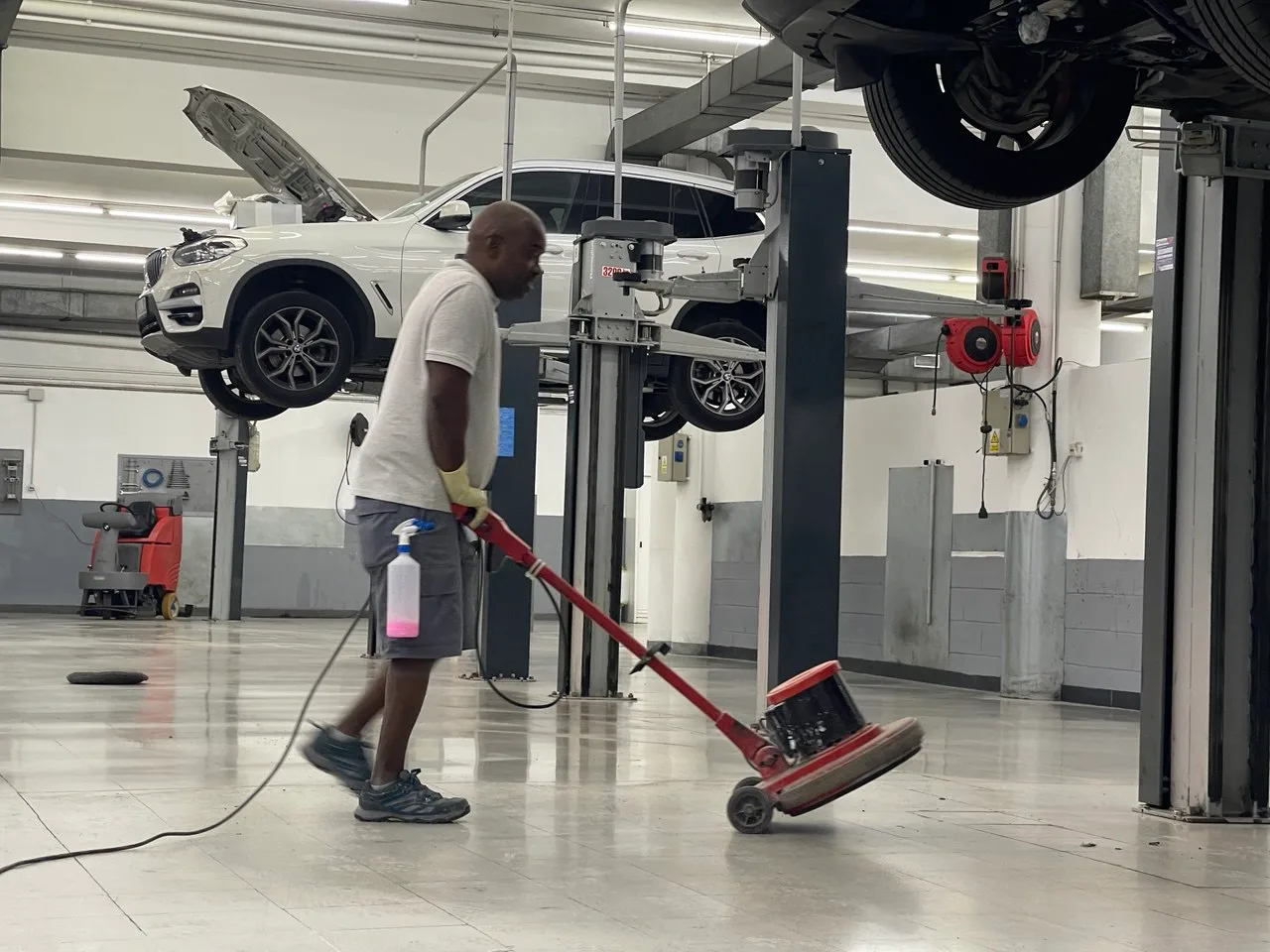 Empleado de Servicios24 puliendo el suelo del taller del concesionario BMW de Lleida, con un vehículo elevado al fondo