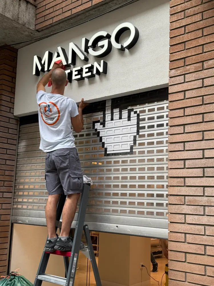 Empleado de Servicios24 limpiando la persiana de una tienda Mango subido a una escalera